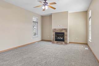 Bright, empty living room with neutral carpet, tiled fireplace, ceiling fan, and tall windows with blinds.
