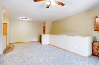 A carpeted living room with a ceiling fan and a half wall separating the stairs down to the door.