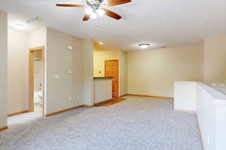 A carpeted living room with a ceiling fan and a half wall separating the stairs down to the door.