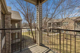 Covered apartment balcony with metal railing overlooking mature trees, landscaped grounds, and neighboring community buildings.