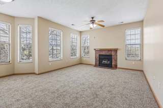 Bright apartment living room with a corner fireplace, carpeted flooring, ceiling fan, and six tall windows with blinds.