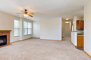 A living room with a fireplace, several tall windows, a ceiling fan, and an opening to the kitchen and hallway.