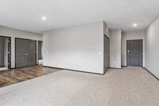 Spacious apartment living room with plush carpeting, recessed lighting, and an open layout connecting to the kitchen and entryway.