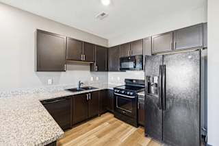 Modern apartment kitchen with granite-look countertops, dark cabinetry, black oven and refrigerator, and wood-style flooring.