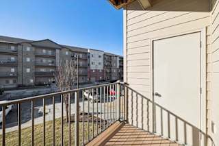 A balcony with a white door and a metal railing overlooks a parking lot and apartment buildings.