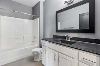 Black-framed mirror and speckled dark countertop contrast with a white vanity and tub-shower combo in this apartment bathroom.