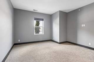 Neutral apartment bedroom with soft gray walls, beige carpeting, dark baseboards, and a single window overlooking trees.