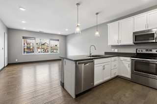 Open-concept apartment kitchen flows into a sunlit living area, with white cabinetry, dark counters, pendant lights, and stainless steel appliances.