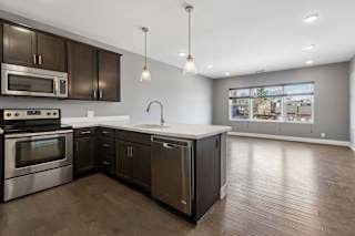 Dark cabinetry and a white peninsula define this open apartment kitchen, flowing into a living area with wide windows and wood-look floors.