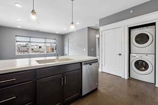 Stacked in-home laundry sits just off the kitchen, beside a dark cabinet island with a deep sink, dishwasher, and pendant lights.