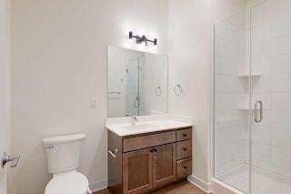 A modern bathroom featuring a white toilet, a wooden vanity with a large mirror, and a glass-enclosed shower. The space is bright with clean lines.