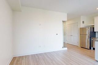 Bright, minimalist room with white walls and light wood floors. A stainless steel fridge stands in the corner, adjacent to a double door and kitchen counter.