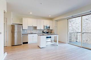 Modern kitchen with white cabinets, stainless steel appliances, and a central island on light wood flooring. Large glass doors lead to a balcony.