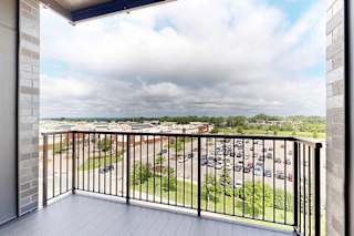 A balcony with a black railing overlooks a parking lot and greenery.