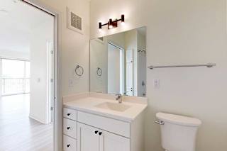 Modern bathroom featuring a white vanity, large mirror, chrome fixtures, and a toilet. Natural light streams in from the adjacent room.