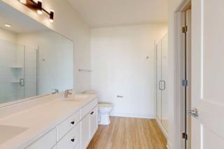 Modern bathroom with white cabinets, a large mirror, single sink, toilet, glass shower enclosure, and wood-look flooring.