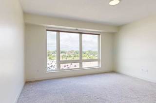 Bright, empty beroom with light-colored walls and carpet, featuring a large window that offers a view of greenery and parking below.