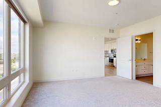 Bright, empty bedroom with beige carpet and a large window. Open doors reveal a modern kitchen and bathroom, creating a clean, inviting atmosphere.
