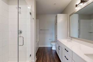 Modern bathroom with a glass shower on the left, wooden floors, white vanity with a large mirror, and a toilet in the background.