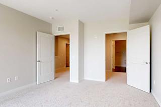 A bright, empty room with light gray walls and beige carpet has two open white doors leading to a hallway and closet.