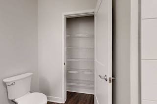 Bathroom with white toilet on the left, adjacent to an open door revealing a pantry with empty wire shelves. Walls are light gray, floor is dark wood.