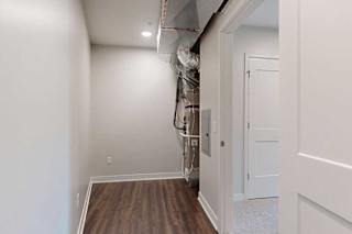 Narrow storage space with hardwood floor and white walls, leading to a utility area with exposed ductwork and piping. A door to the right opens to a carpeted room.