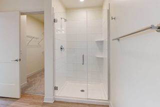 Modern bathroom with a glass-enclosed shower featuring white tiles, chrome fixtures, and a metal towel bar. Open door reveals a carpeted walk-in closet.