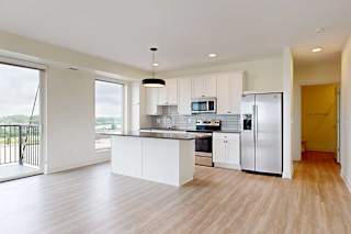 Modern, bright kitchen with light wood flooring, white cabinets, and stainless steel appliances. Large windows and a small balcony offer an open view.