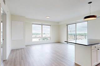 Modern, unfurnished apartment with light wood flooring, large windows showing a city view, white walls, and a black pendant light over a kitchen island.
