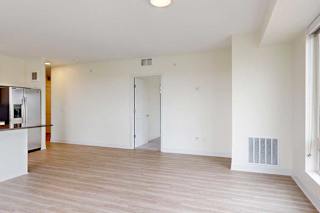 Minimalist, unfurnished apartment with light wood flooring. Features a modern kitchen on the left, a central door, and large windows on the right.