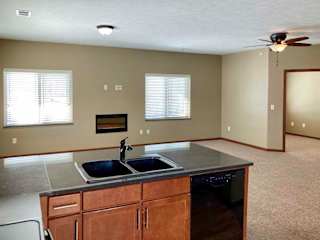 A kitchen peninsula counter with a sink and dishwasher that faces a living room with large windows.