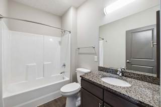Apartment bathroom with a granite-look single-sink vanity, dark cabinetry, a large mirror, and a tub-shower combination with a white surround and modern fixtures.