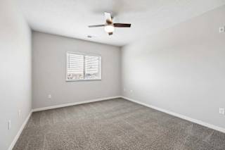 Spacious apartment bedroom with carpeted flooring, a ceiling fan, neutral walls, and a window with blinds providing natural light.