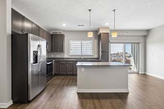 Open-concept apartment kitchen with a large island, dark cabinetry, granite-look countertops, stainless steel appliances, pendant lighting, and a spacious living area with a sliding glass door leading to a private balcony.