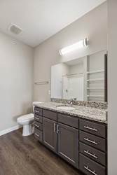 Apartment bathroom with a granite-look single-sink vanity, dark cabinetry, a large mirror with overhead lighting, and wood-style flooring.