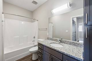 Apartment bathroom with a granite-look single-sink vanity, dark cabinetry, a large mirror, and a tub-shower combination with a white surround.