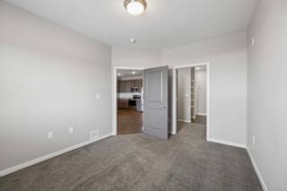 Spacious apartment bedroom with carpeted flooring, neutral walls, and an open layout featuring a walk-in closet and hallway access through gray interior doors.