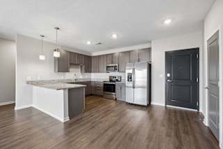 Open-concept apartment kitchen with a large island, dark cabinetry, granite-look countertops, stainless steel appliances, pendant lighting, and wood-style flooring.
