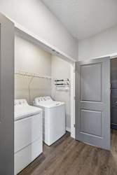 Apartment laundry room with side-by-side washer and dryer, built-in wire shelving, wood-style flooring, and a gray interior door leading to the hallway.