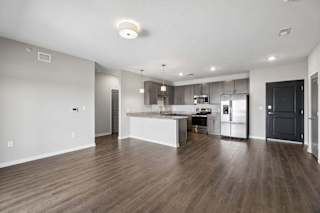 Spacious open-concept apartment living area with wood-style flooring, a modern kitchen featuring a large island, dark cabinetry, stainless steel appliances, and recessed lighting.