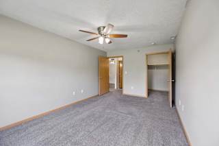 Spacious apartment bedroom with soft carpeting, a ceiling fan, an open closet, and access to an adjoining hallway.