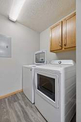 Dedicated apartment laundry room with a side-by-side washer and dryer, overhead storage cabinets, and wood-style flooring.