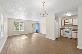 A well-lit, empty room with a chandelier and a kitchenette in the background.