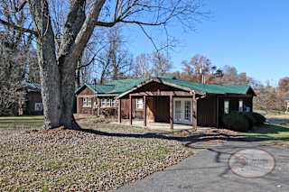 A rustic two bedroom cabin style home with green roof and paved driveway.