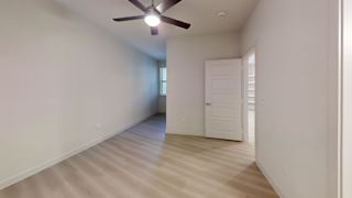 A room with a ceiling fan and light, wooden flooring, and a door at the end of the hallway.