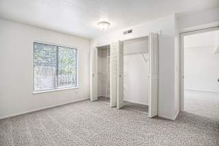 A bedroom with white walls and a carpeted floor with a window and open closet doors at Canal Club Apartments in Lansing, MI