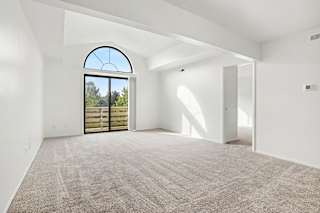 A large empty living room with a carpeted floor and a large arched window at Pine Knoll Apartments, Battle Creek