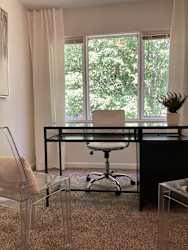 A black desk with a glass top and a white chair in front of a window with trees visible outside.