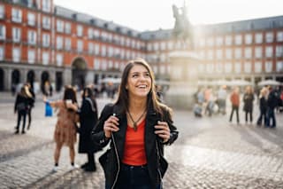 A woman in a red top and black jacket is standing in front of a building with a fountain in the background.