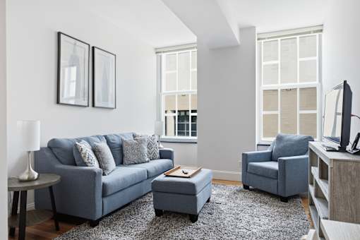 A living room with a grey couch, a grey armchair, a grey ottoman, a grey rug, a grey coffee table, a grey lamp, a grey television, and a grey wall.